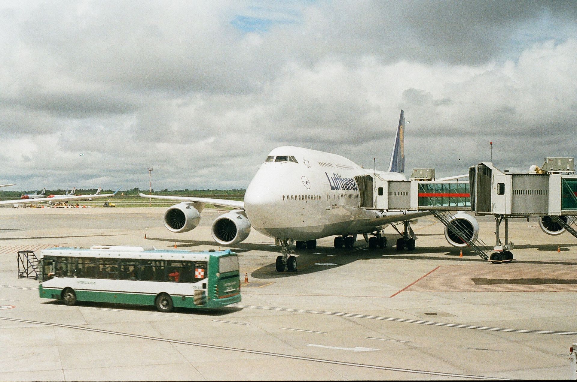 A large airplane docked at an airport gate with a shuttle bus nearby under cloudy skies.