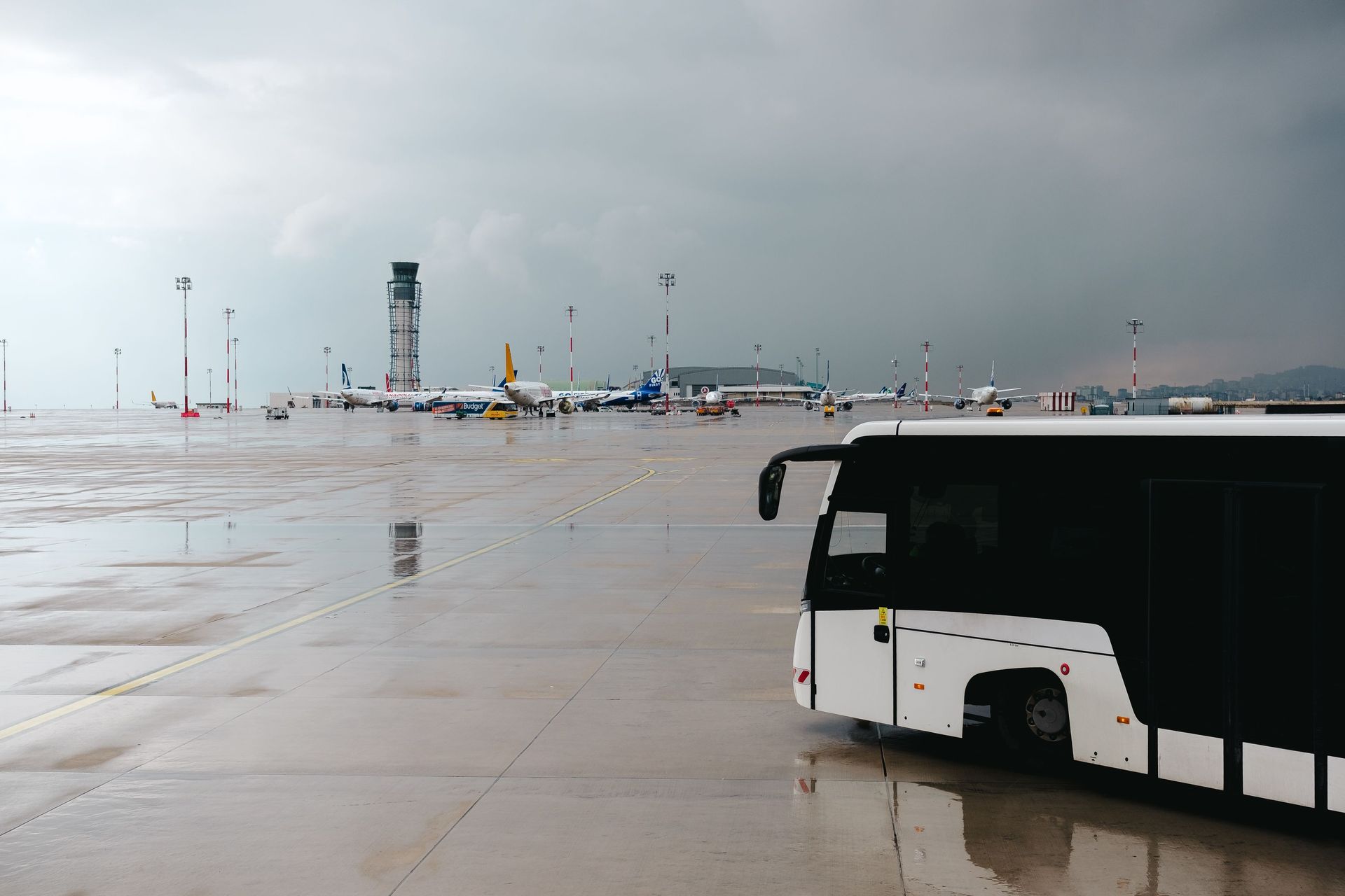 Rainy airport tarmac featuring a passenger bus with airplanes in the background.